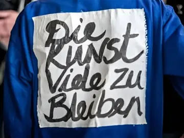 Rückansicht einer Person in blauer Kleidung. Auf dem Rücken ist ein weißes Stoffstück befestigt, auf dem handschriftlich in schwarzer Farbe steht: „Die Kunst Viele zu Bleiben“. Der Schriftzug wirkt wie ein künstlerisches Statement oder Protestausdruck. Im Hintergrund sind unscharf weitere Menschen zu erkennen.