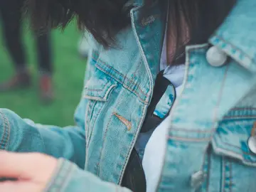 **Alternativtext (Barrierefreiheit):** Nahaufnahme einer Person mit dunklem Haar in einer hellblauen Jeansjacke. In der Brusttasche steckt eine Sonnenbrille, in deren Gläsern sich der Himmel mit Wolken spiegelt. Im Hintergrund ist unscharf eine grüne Wiese und eine weitere Person zu erkennen.
