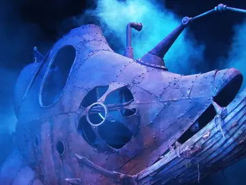 Ein großes, fantasievoll gestaltetes U-Boot in Form eines Fischkopfes steht auf einer Bühne, umgeben von blauem Nebellicht. Das Gefährt besteht aus rostigem, genietetem Metall und erinnert an ein Tiefseeungeheuer mit Bootsrumpf-Maul, Bullaugen und einem mechanischen Arm mit Laterne. Die Szene wirkt geheimnisvoll und märchenhaft, als stamme sie aus einer Unterwasserwelt.