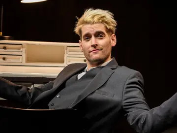 Ein junger Mann mit blondem Haar sitzt selbstbewusst in einem Anzug vor einem weißen Schreibtisch und schaut mit leicht spöttischem Blick in die Kamera. Er hebt beide Arme seitlich mit offenen Händen, als wolle er sagen: „Was soll ich machen?“ Eine Schreibtischlampe leuchtet von oben, im Hintergrund ist es dunkel.