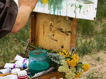 Nahaufnahme einer Hand, die an einem Landschaftsbild in Aquarellfarben auf einer Staffelei arbeitet. Daneben ein Holzkasten mit Malutensilien, einem Glas mit grüner Farbe und frischen gelben Blüten.