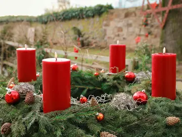 Ein Adventskranz aus Tannenzweigen, geschmückt mit roten Kugeln, Tannenzapfen und vier großen roten Kerzen, von denen zwei angezündet sind.