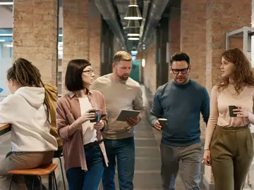 Fünf Personen gehen gemeinsam durch einen Büroflur, einige halten Kaffeebecher in der Hand, während sie sich unterhalten.