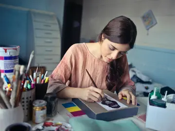 Eine junge Frau sitzt an einem Tisch und malt konzentriert ein Bild auf einer Leinwand. Auf dem Tisch liegen viele Mal- und Zeichenutensilien wie Pinsel, Stifte und Farben.