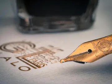 Eine Nahaufnahme einer Tintenfeder mit einem Tropfen Tinte auf Papier, im Hintergrund ein Glas Tintenfläschchen.