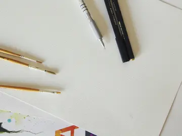 Leeres Blatt Papier auf einem Tisch, daneben drei Malpinsel, ein schwarzer Stift und ein silberner Zeichenstift.