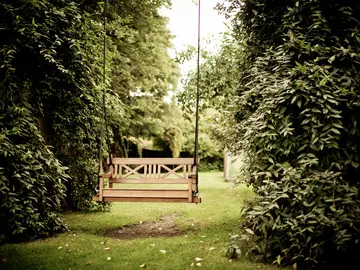Eine hölzerne Gartenschaukel hängt an dicken Seilen zwischen üppig grünen Büschen und Bäumen.