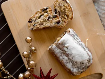 Ein angeschnittenes Stück Christstollen liegt auf einem Holzbrett, bestäubt mit Puderzucker. Neben dem Gebäck befinden sich weihnachtliche Dekorationen und Lichterketten.