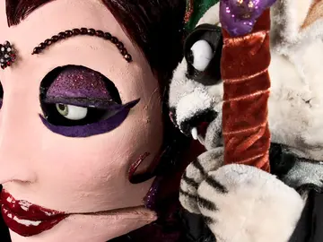 Detailaufnahme einer glamourösen Puppenfigur mit glitzerndem violetten Augen-Make-up und rotem Lippenstift. Neben ihrem Gesicht lugt ein kleines weißes Tier mit spitzen Zähnen und schwarzem Umhang hervor.