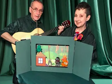 Zwei Personen stehen vor grünen Bühnenvorhängen. Links spielt ein Mann Gitarre, rechts hält eine Frau eine kleine Stabpuppe in Form eines bunten Hahns. Vor ihnen steht ein kleines Tischtheater mit aufgeklappter grüner Kulisse. In der Mitte ist ein Fenster mit Figuren: ein Frosch sitzt auf dem Dach eines kleinen Hauses, daneben stehen eine Maus, ein Wolf und ein Fuchs als Puppen. Die Szene zeigt das mobile Wohnzimmertheaterstück „Das Tierhäuschen“.