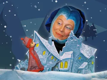 Ruth Brockhausen als Frau Mond trägt ein blaues Kostüm und schaut lächelnd auf eine kleine Puppenfigur in rotem Mantel und Mütze. Die Puppe steht vor einer hellblauen, stilisierten Häuserkulisse im Schnee. Der Hintergrund zeigt einen dunklen Winterhimmel mit Schneeflocken und Silhouetten von Tannen.