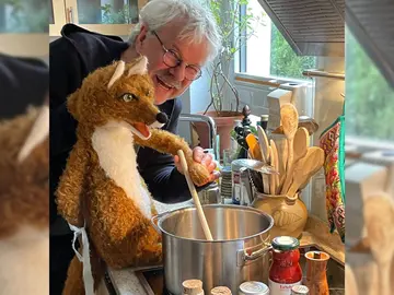 Ein älterer Mann mit grauen Haaren und Brille steht in einer Küche und hält eine Fuchs-Handpuppe. Gemeinsam rühren sie mit einem Holzlöffel in einem großen Kochtopf auf dem Herd. Auf der Arbeitsfläche stehen Kochutensilien, Gläser und ein Topf mit Holzlöffeln. Der Mann lächelt, die Szene wirkt spielerisch und humorvoll.