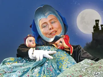 Ruth Brockhausen als Frau Mond sitzt vor dem Nachthimmel. Sie trägt einen blauen Helm und hält zwei Puppen im Arm: links die Prinzessin mit Krönchen, rechts den kleinen Hofnarren in rotem Kostüm. Im Hintergrund steht eine dunkle Burg vor dem hellen Vollmond.
