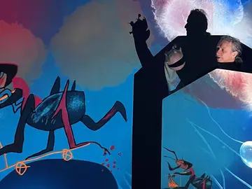 Ein Performer steht als dunkle Silhouette vor zwei leuchtenden Leinwänden mit farbenfrohen animierten Illustrationen – links fantasievolle Vogelwesen, rechts eine blaue Unterwasserwelt mit Ameisen und einer Seifenblase. In der Hand hält er eine kleine Figur.