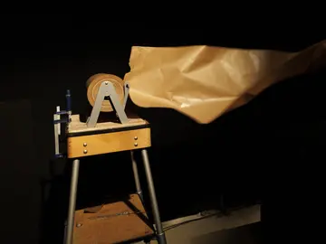 Eine Performerin in schwarzem Kostüm reißt ein Stück Packpapier von einer Rolle ab, die auf einem Holzgestell steht. Eine Performerin in schwarzem Kostüm reißt ein Stück Packpapier von einer Rolle ab, die auf einem Holzgestell steht. Die Bewegung ist unscharf, der Hintergrund schwarz. Theaterfoto.