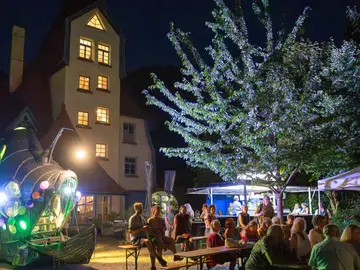 Abendliche Freiluftveranstaltung vor dem Theater der Nacht. Links eine bunt beleuchtete, futuristische U-Boot-Skulptur mit bunten Leuchtkugeln, daneben ein altes Holzboot. Zahlreiche Besucherinnen und Besucher sitzen auf Bierbänken oder stehen zusammen, im Hintergrund ein Pavillon und ein beleuchteter Baum. 