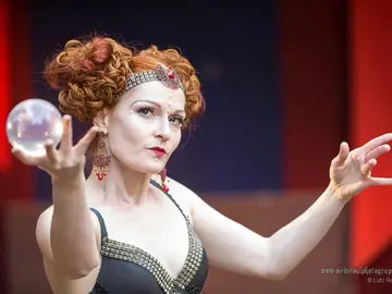 Contact-Juggling-Künstlerin mit rotem Hochsteckfrisur, Stirnschmuck und roten Ohrhängern lässt eine gläserne Kristallkugel über ihrer Hand schweben, beide Arme ausgestreckt; im Hintergrund verschwommene rot-blaue Bühnenbeleuchtung.