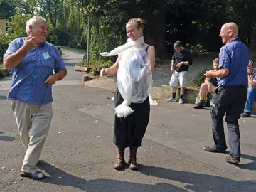 Freies Workshop-Training im Freien: Eine lachende junge Frau trägt eine improvisierte Figur aus durchsichtiger Plastikfolie um Hals und Oberkörper, die zwei Männer mit Fäden von außen führen; im Hintergrund sitzen und stehen weitere Workshop-Teilnehmende lachend auf einem Weg zwischen Bäumen.