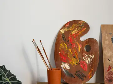 Eine mit Farbresten bedeckte Holzpalette und mehrere Pinsel stehen auf einem Holztisch vor einer weißen Wand.