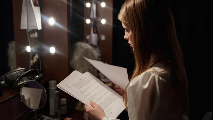 Eine junge Frau steht vor einem beleuchteten Spiegel, in den Händen hält sie mehrere Textblätter.