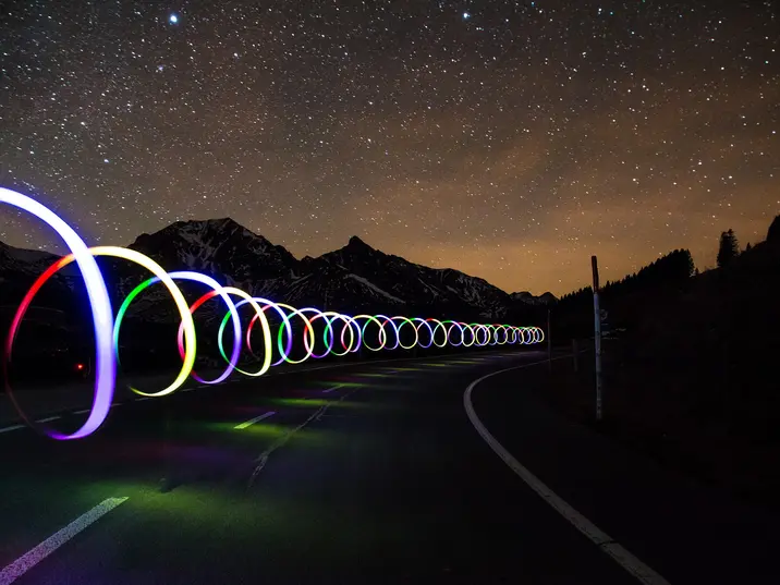 Eine Straße bei nacht mit Sternenhimmel. Über der Straße "schweben" Lichtzeichnungen: eine spiralförmige farbige Lichtröhre