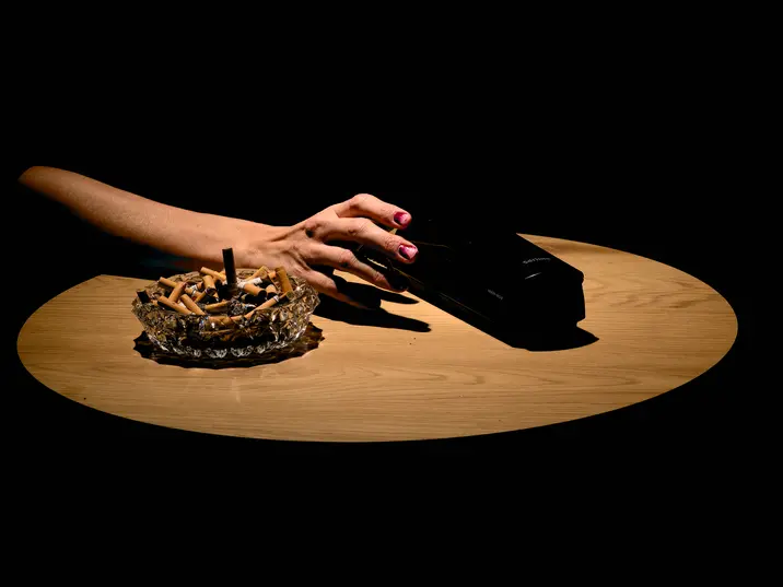Ein Tisch, darauf ein voller Aschenbecher, von der linken Seite greift eine Hand nach einem Telefonhörer.
