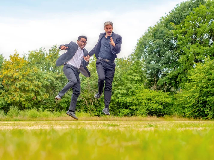 Zwei Männer im Anzug springen in die Luft, im Hintergrund grüne Landschaft.