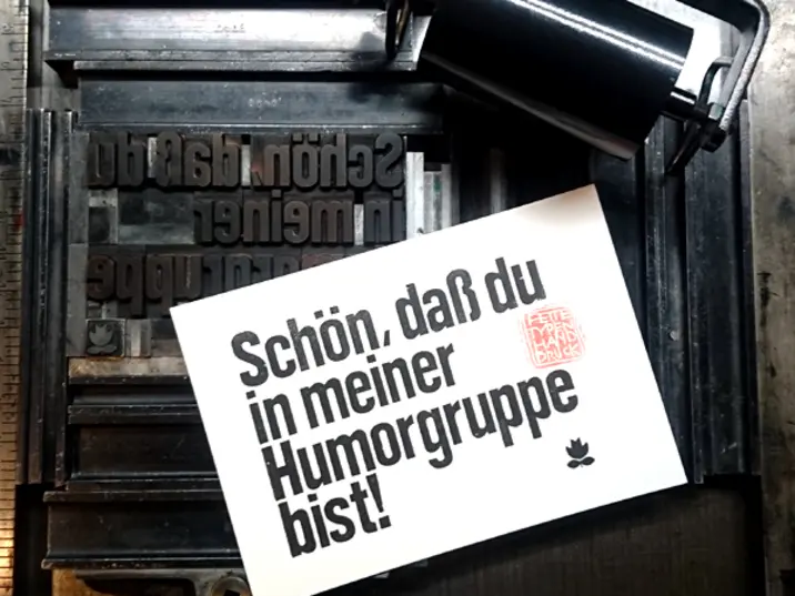eine schwarze Druckmaschine mit Rolle. Auf einer weißen Karte steht in schwarzer Tinte gedruckt: "Schön, daß du in meiner Humorgruppe bist!"