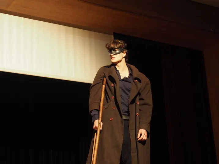 Ein Mann mit Maske / Augenbinde, er trägt eine Art Militärmantel und hstceinecaltmidische Gehhilfe.