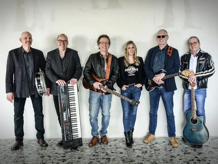 Die Band Rocktail mit 6 Mitgliedern, davon 5 Männer und eine Frau stehen mit ihren Instrumenten in Reihe. Sie tragen alle Jeans und schwarze Jacken.