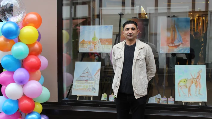 Der Künstler Abdulkarim Alhasan steht vor einem Fenster mit Bildern. Neben ihm sind bunte Luftballons