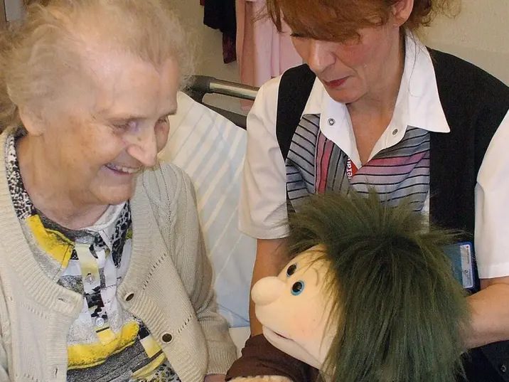 Eine jüngere Frau sitzt neben einer Seniorin und beide schauen auf eine Puppe
