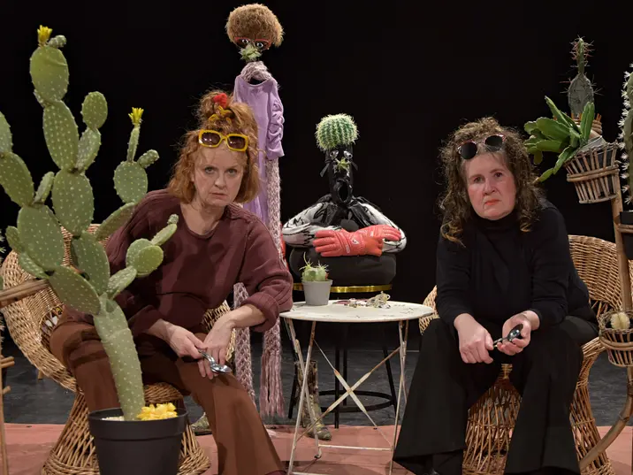 Zwei Frauen sitzen in Korbstühlen auf einer Theaterbühne, umgeben von zahlreichen Kakteen in Töpfen und Pflanzenständern. Beide tragen Sonnenbrillen im Haar und halten kleine metallische Gegenstände in den Händen. Im Hintergrund steht eine skurrile Figur mit einem Kaktus als Kopf und roten Boxhandschuhen. Die Szene wirkt humorvoll und absurd, wie ein Teil einer Theaterperformance.