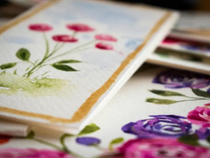 Aquarellgemälde mehrerer Blumen auf strukturiertem Papier, übereinander liegend. Die vorderen Bilder zeigen zart gemalte rosa, lila und rote Blüten mit grünen Blättern, teilweise von einem goldenen Rahmen umgeben. Die Darstellung ist künstlerisch und unscharf im Hintergrund.