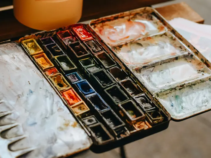 Nahaufnahme einer gebrauchten Aquarellfarbpalette mit vielen unterschiedlich gefüllten Farbnäpfen. Die Farben sind leicht vermischt, und einige Bereiche der Palette zeigen getrocknete Farbspuren und Wasserreste.