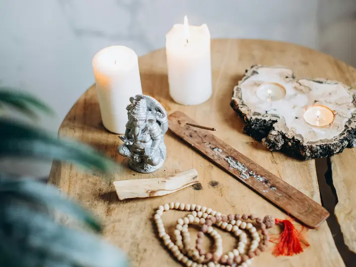 Ein kleiner runder Holztisch mit brennenden Kerzen, einer Ganesha-Statue, Räucherstäbchenhalter, Holzperlenketten mit roten Quasten und einem Stück Palo Santo.