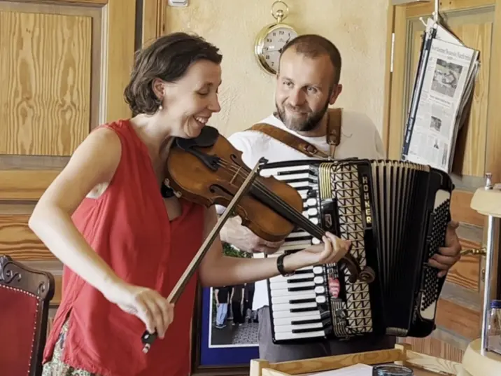 Zwei Musiker*innen spielen Geige und Akkordeon mit Freude in einem gemütlichen Raum mit Holzdekor.