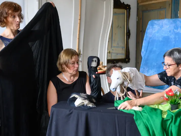 Vier Erwachsene gestalten ein Puppenspiel mit fantasievollen Figuren an einem Tisch, umgeben von Tüchern und Requisiten.
