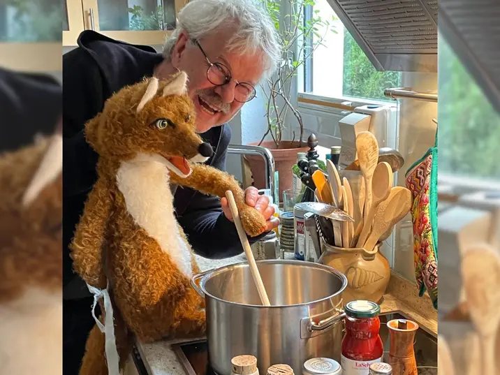 Ein älterer Mann mit grauen Haaren und Brille steht in einer Küche und hält eine Fuchs-Handpuppe. Gemeinsam rühren sie mit einem Holzlöffel in einem großen Kochtopf auf dem Herd. Auf der Arbeitsfläche stehen Kochutensilien, Gläser und ein Topf mit Holzlöffeln. Der Mann lächelt, die Szene wirkt spielerisch und humorvoll.