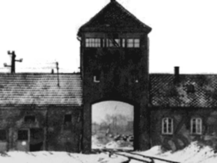 Historische Schwarz-Weiß-Aufnahme des Lagerkomplexes Auschwitz: Zu sehen ist eine lange, von einem zentralen Wachturm überragte Gebäudezeile mit einem großen Tordurchgang. Entlang der Gebäude verläuft eine Mauer mit mehreren Fenstern und Eingängen. Im Vordergrund liegt Schnee.